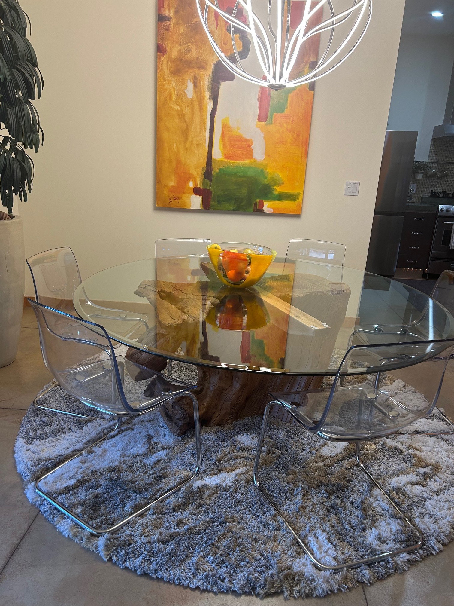 A dining room with a glass-top table supported by a natural wood base, surrounded by five transparent plastic chairs with chrome legs. On the table is a clear glass bowl filled with oranges, lemons, and a green apple. The table sits on a plush, circu