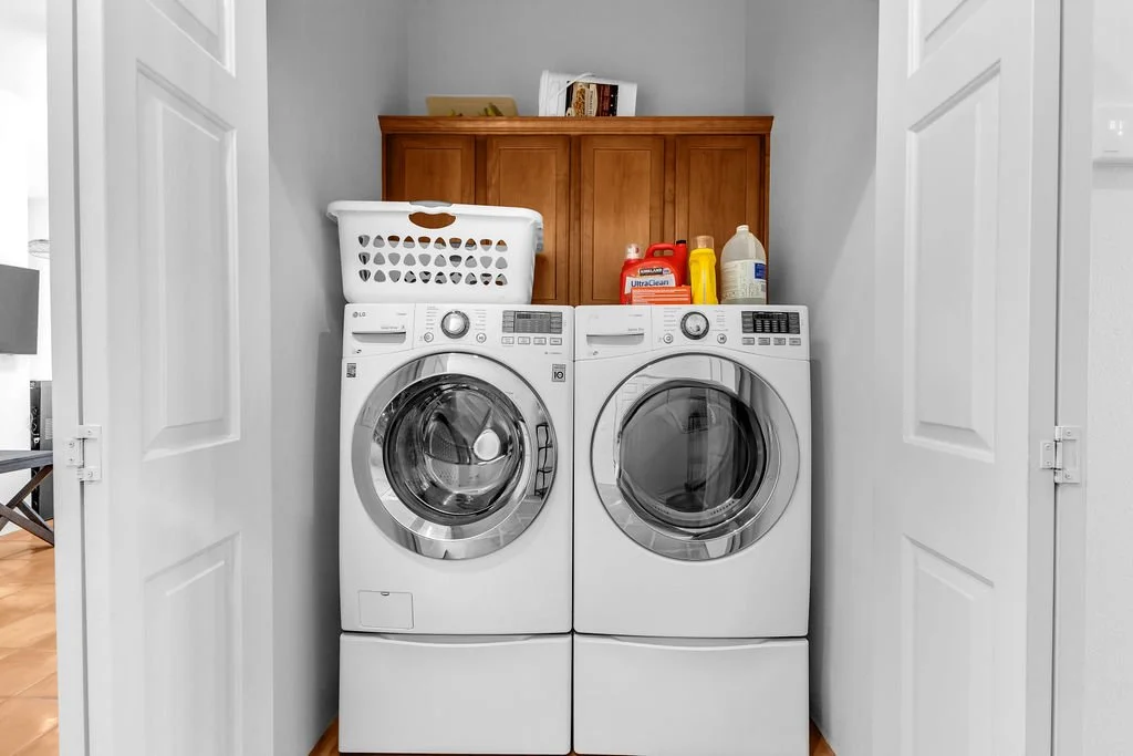 A front-loading washing machine and dryer in a laundry closet, with laundry supplies on top, framed by white double doors.