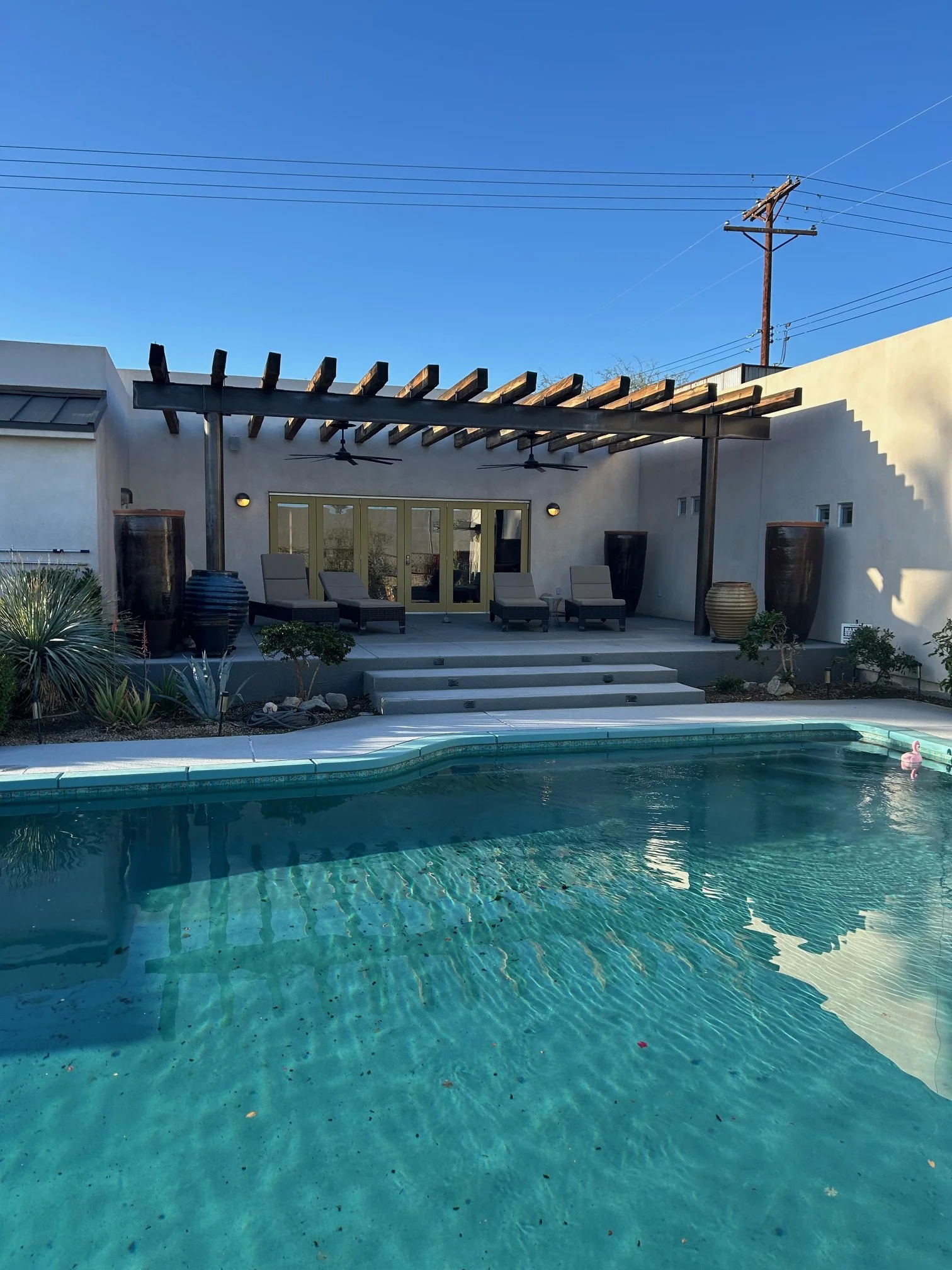 Backyard pool with clear water and a floating pink flamingo pool float, surrounded by plants, lounge chairs, a cabana, and a blue sky with power lines overhead.