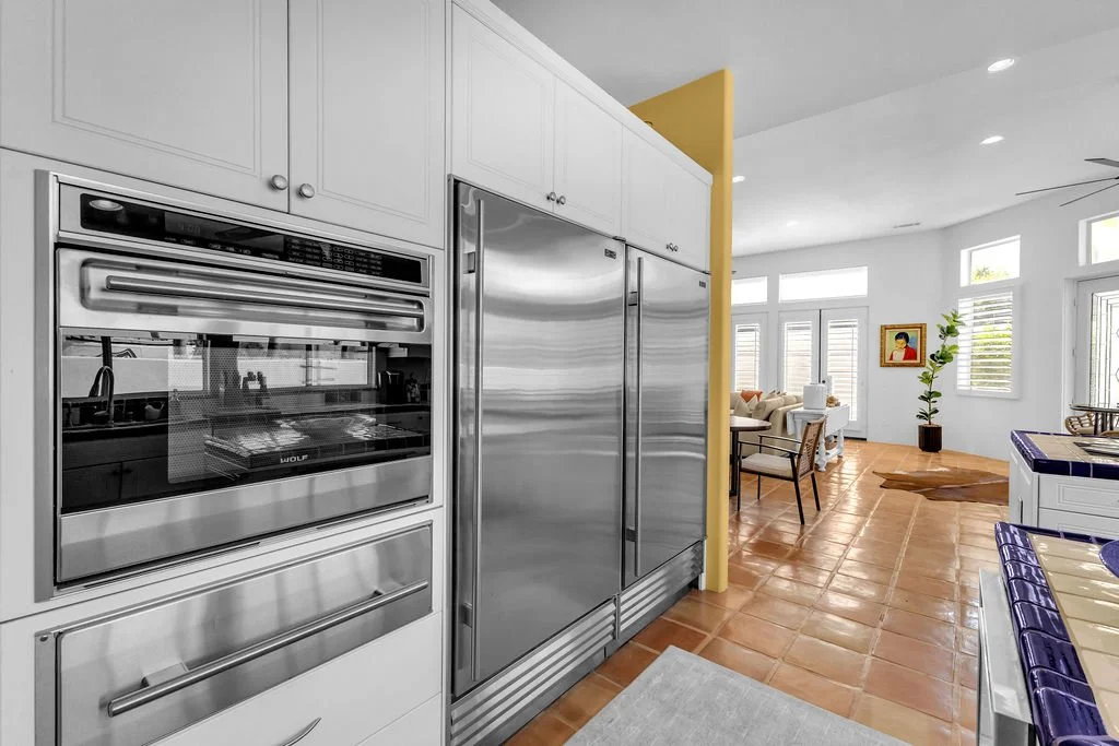 Modern kitchen with stainless steel appliances and terracotta tile flooring, opening to a bright dining and living area with large windows and a potted plant.
