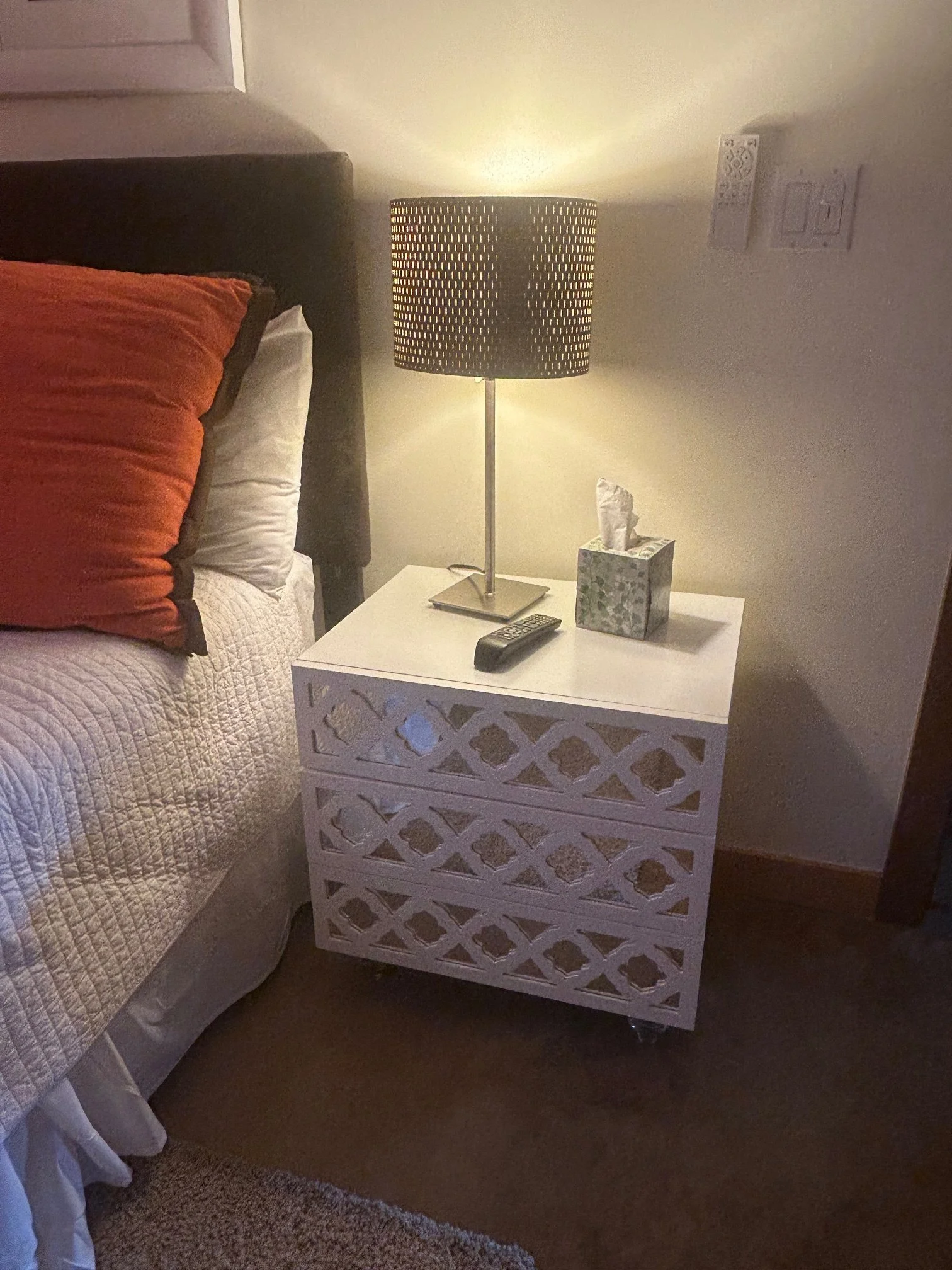 A bedroom nightstand with a black lamp, tissue box, and remote control next to a bed with orange and white bedding.