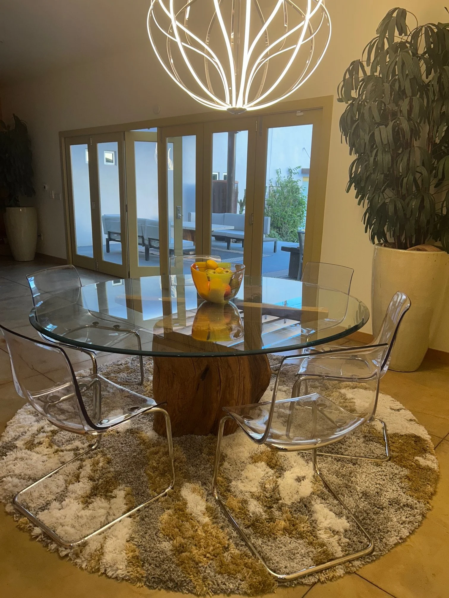 A modern dining room with a glass-top table surrounded by transparent acrylic chairs, a large shaggy rug underneath, a chandelier hanging from the ceiling, and sliding glass doors leading to an outdoor patio with seating and greenery.