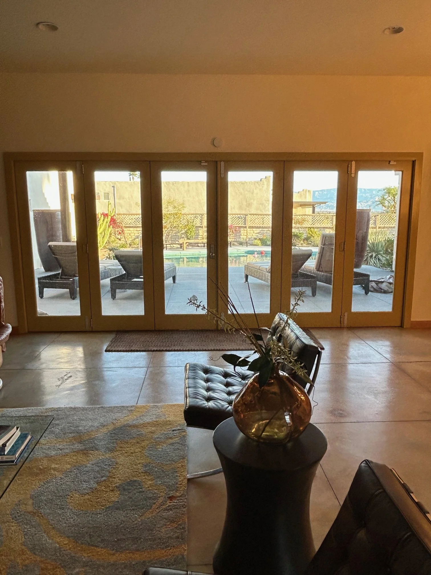 View of an indoor living space with a large sliding glass door leading to an outdoor patio with chairs, plants, and mountains in the background. Inside, there is a vase with dried branches on a small round table, a patterned area rug, and a few chair
