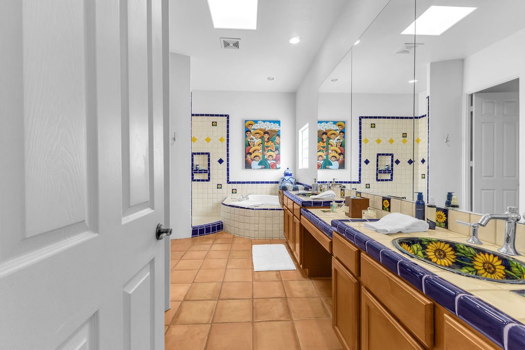 A bathroom featuring a double sink vanity with sunflower decorated bowls and purple tile accents, a bathtub with a colorful mural above, and white walls with blue and yellow tile patterning.