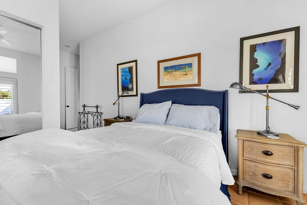Bedroom with white walls, a bed with a blue headboard, white bedding, and three framed art pieces on the wall. There are wooden nightstands with lamps and a mirror reflecting part of the room.