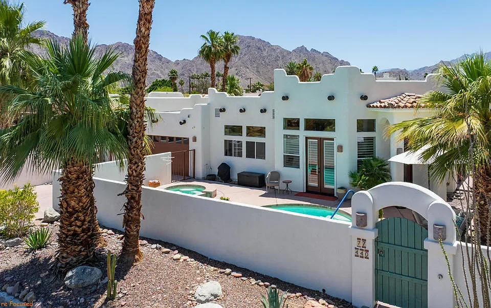 View of a white southwestern-style house with a courtyard, palm trees, and a pool in a desert landscape with mountains in the background.