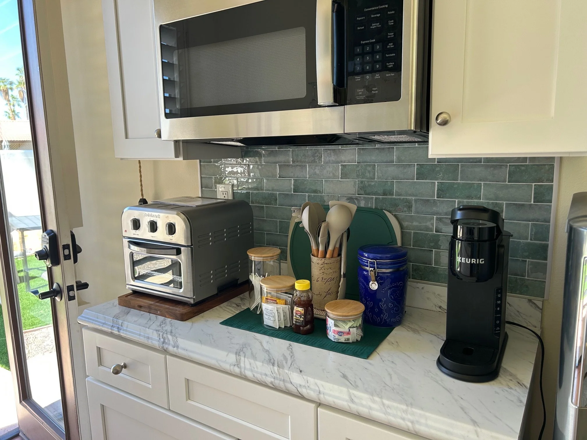 Kitchen counter with a microwave, a small toaster oven, a row of seasonings, a blue thermos, a coffee maker, and various utensils and containers, with a window showing a patio outside.