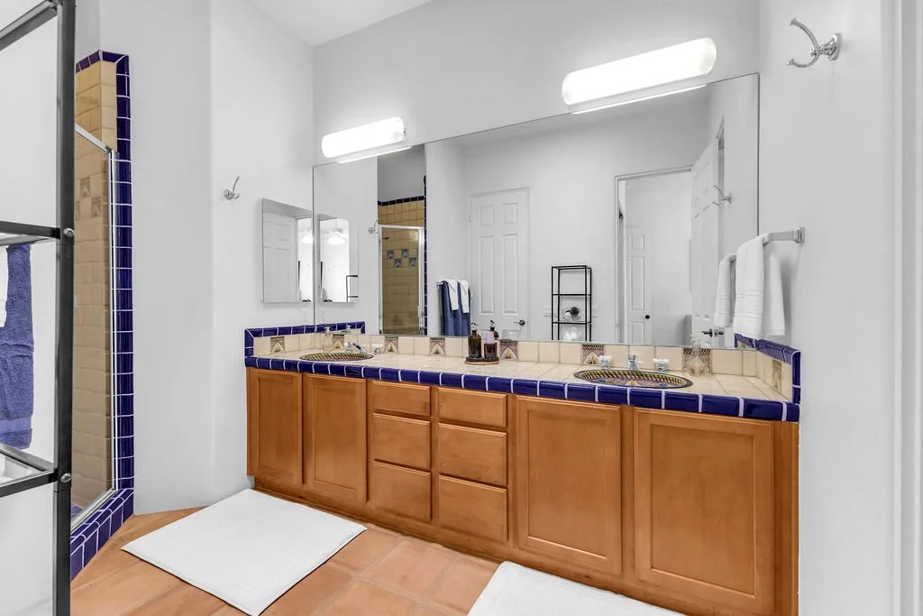Bathroom with double sink vanity, large mirror, blue-tiled countertop, wooden cabinets, white towels, towel rack, and a glass shower enclosure with blue tiles.