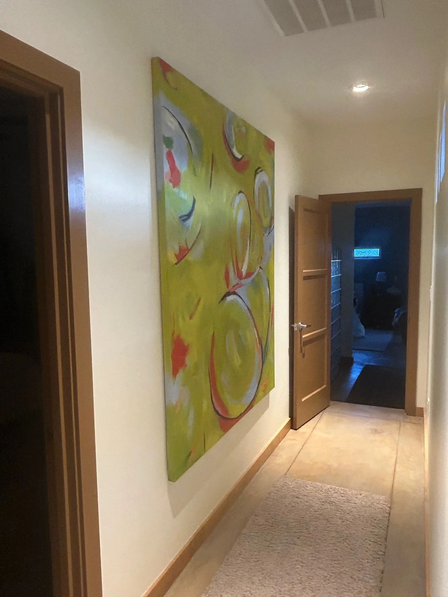 An abstract painting with yellow, red, black, white, blue, and green swirls hanging on a cream-colored wall in a hallway with a beige carpet. There are two wooden door frames, one open, revealing a dark room, and a glimpse of another room with a smal