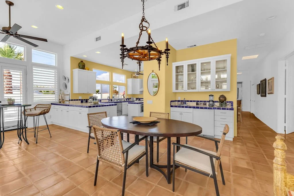 Open kitchen and dining area with white cabinets, yellow accent wall, terracotta tile floor, chandelier, and large windows with white shutters.