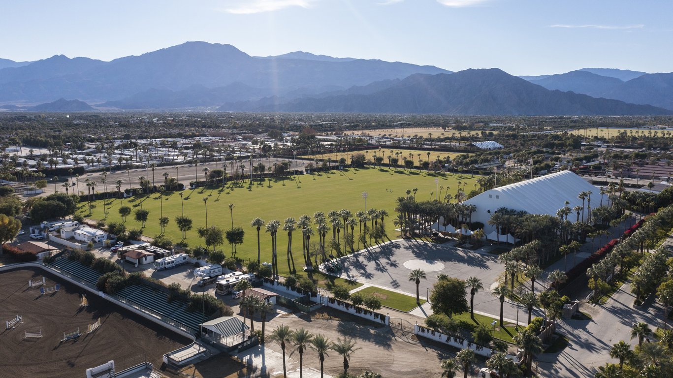 An aerial view of an outdoor sports complex with a large green field, surrounded by tall palm trees, with mountains in the background.
