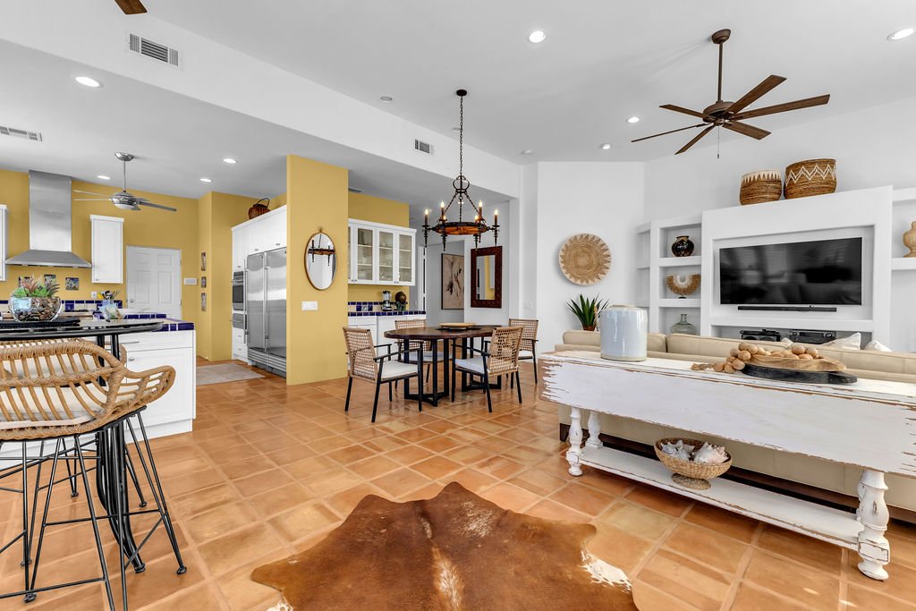 An open-plan living room and kitchen with terracotta tile flooring, white and yellow walls, a ceiling fan, a TV on a white built-in unit, decorative baskets, a chandelier over the dining table, wicker chairs, a cowhide rug, and kitchen appliances.