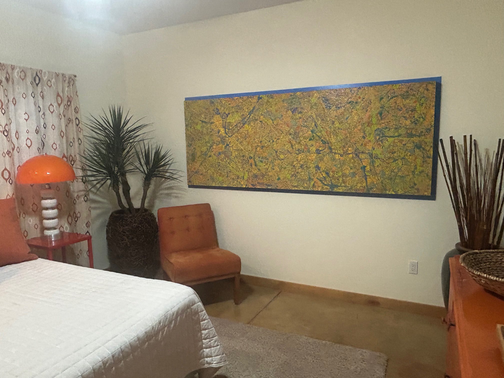 A bedroom corner with a white quilted bed, a vintage orange chair, a small orange side table with a lamp, a potted indoor plant, a large abstract painting on the wall, and a curtain with a geometric pattern.