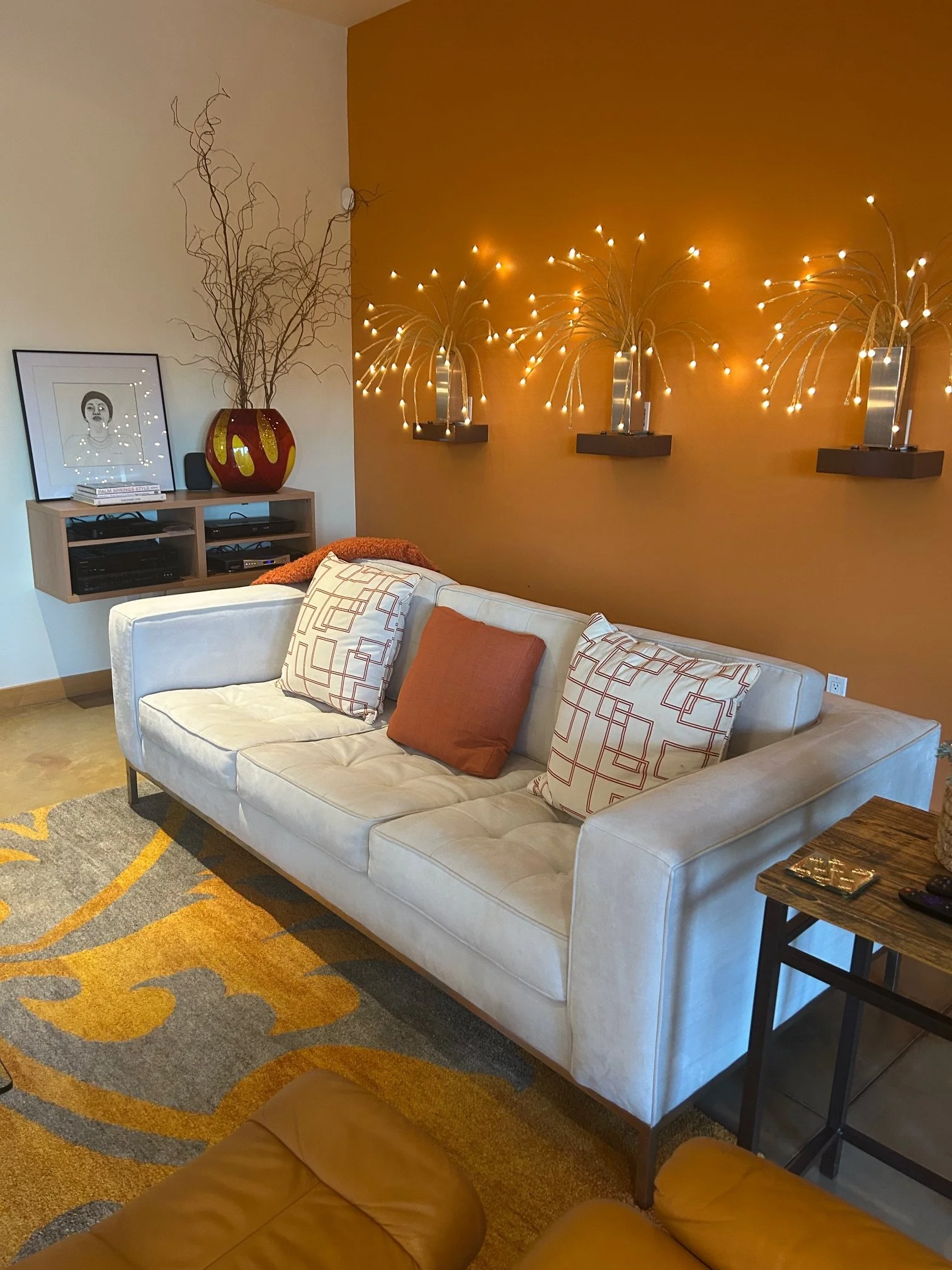 Living room with a white sofa, decorative pillows, a wooden side table, a yellow and gray patterned rug, a wall with a warm orange color, string lights, and a framed black-and-white drawing on a wall-mounted shelf.