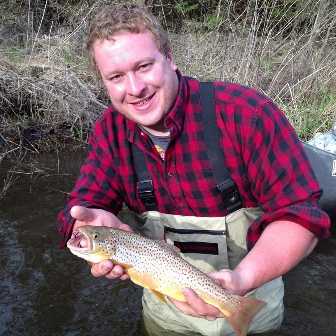 Man in waders holding a caught brown trout in a creek with dry grass and plants in the background.