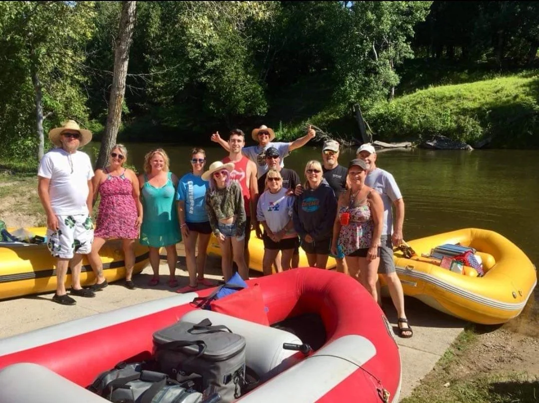 Group of fifteen people standing beside inflatable boats on a riverbank, with trees and greenery in the background.