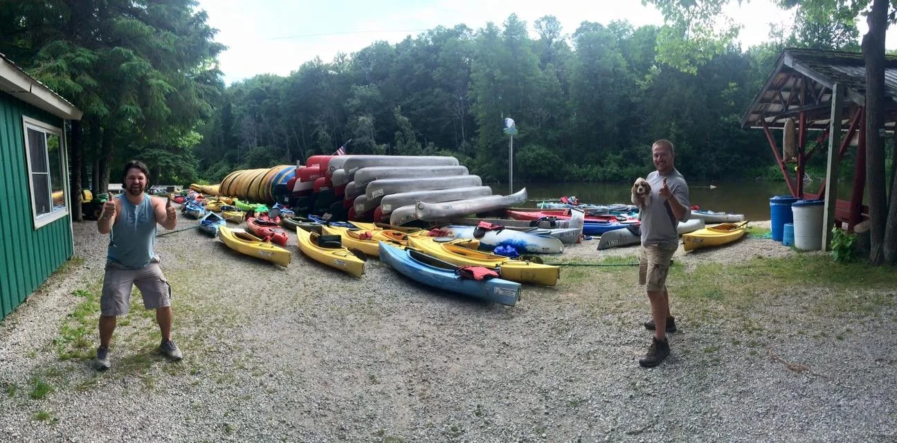Two men stand on a gravel area near the water, smiling and giving thumbs up, with a collection of kayaks, canoes, and paddles stacked on the ground behind them.
