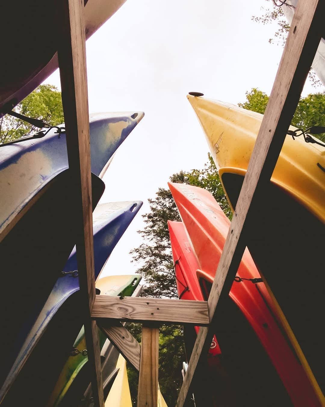 Looking up at colorful kayaks stored on a wooden rack outside, with trees and cloudy sky in the background.
