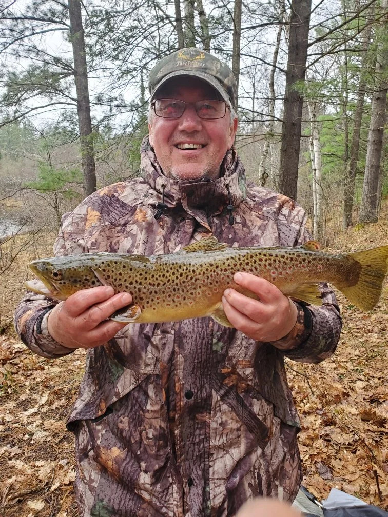 A man in camouflage clothing and a cap smiling and holding a large fish in an outdoor wooded area.