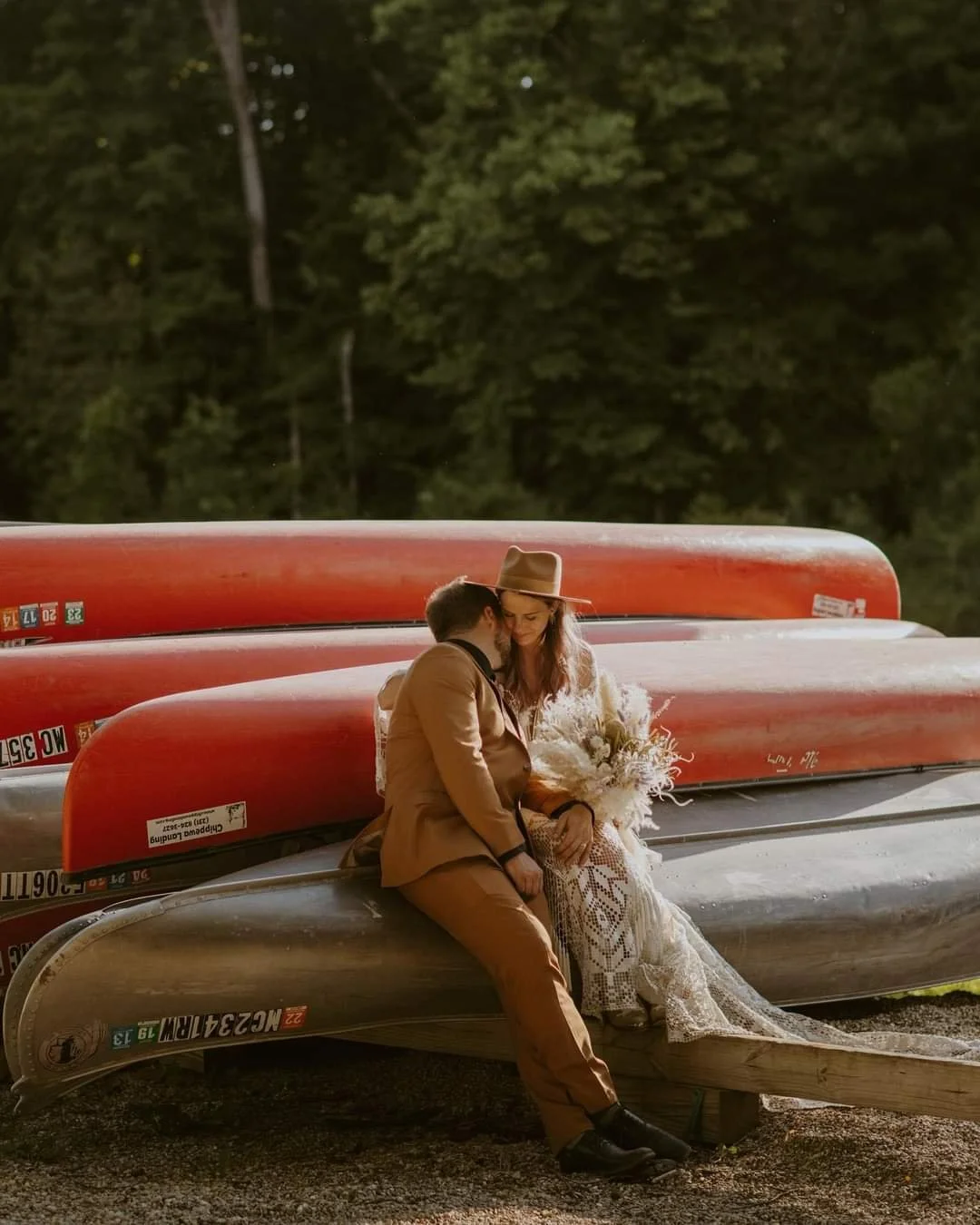 A couple sitting on a silver canoe with red canoes stacked behind them, outdoors at sunset, surrounded by trees, sharing an intimate moment.