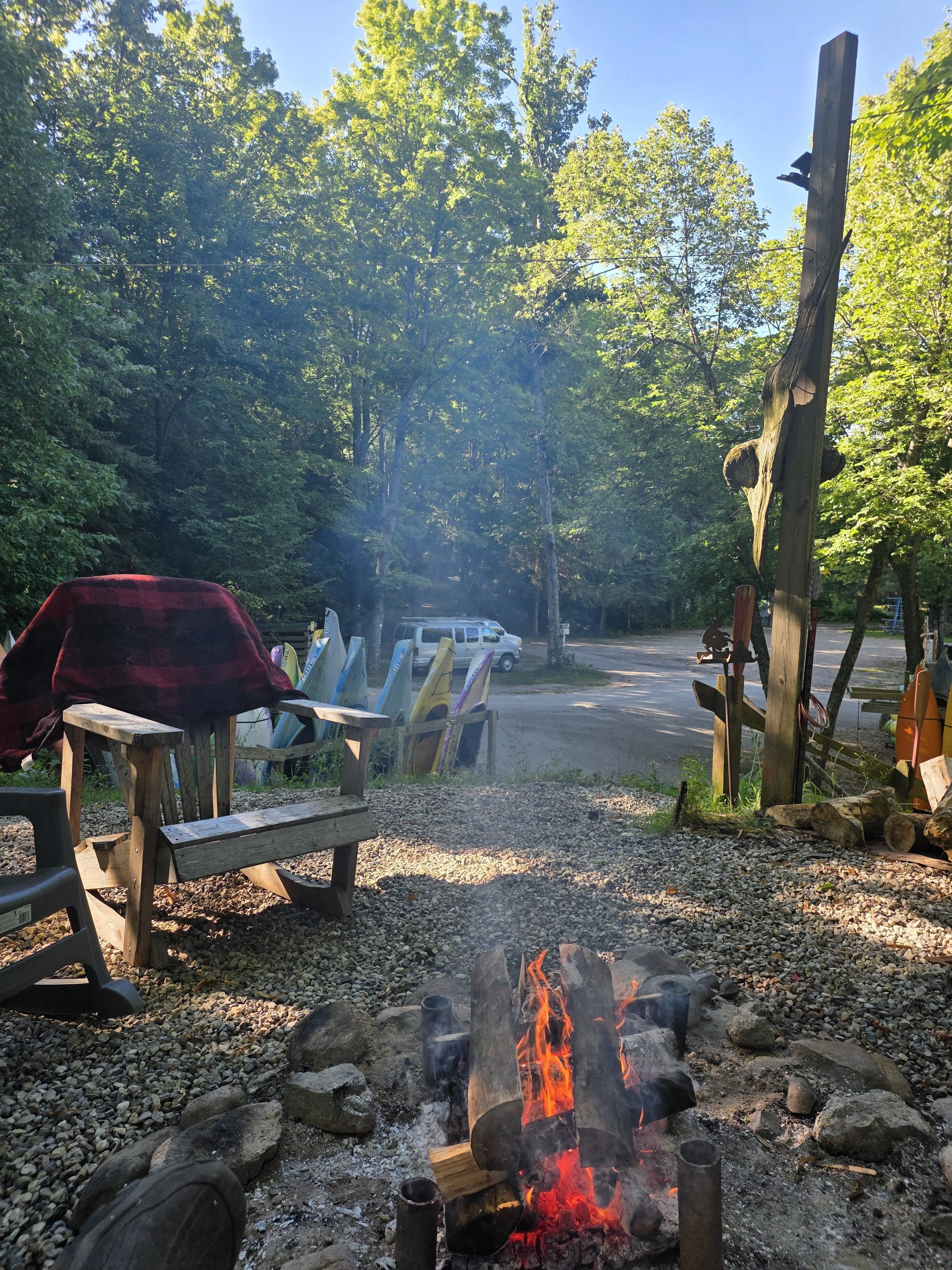 Outdoor scene with a small campfire surrounded by rocks, chairs, and wooden furniture, set in a wooded area with tall trees and a bright blue sky.