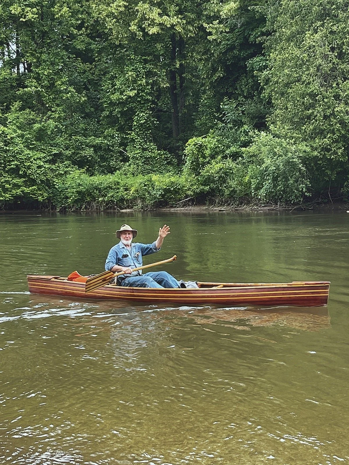 An older man in a blue shirt and hat paddling a wooden canoe on a river, waving with his right hand, with lush green trees in the background.