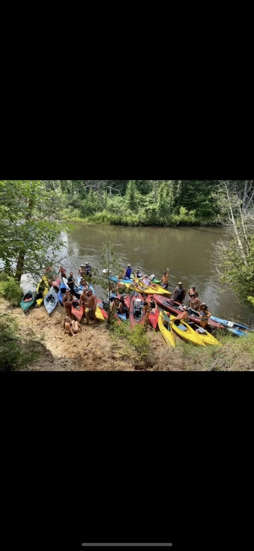 People gather with kayaks on the riverbank before kayaking in a forested area.