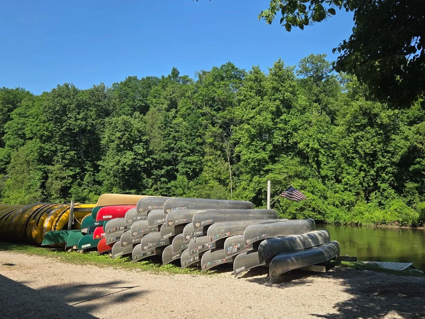 Several canoes and kayaks stacked along a riverbank with lush green trees in the background and an American flag waving nearby under a clear blue sky.