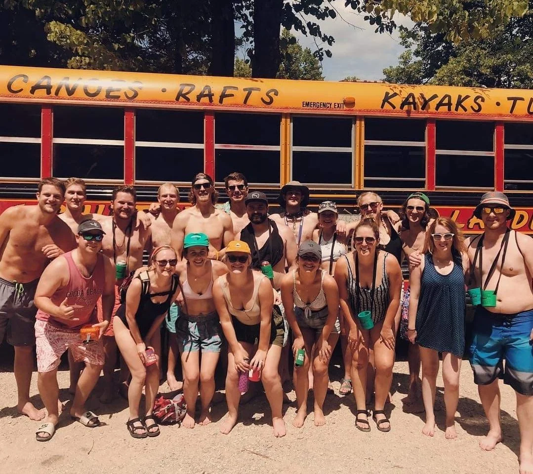 Group of people in swimwear standing in front of a yellow bus labeled 'Canoes, Rafts, Kayaks, Tubes' during daytime at outdoor location.