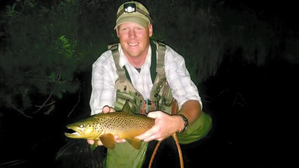 Man in outdoor gear holding a large brown-spotted fish at night in a dark, natural setting.