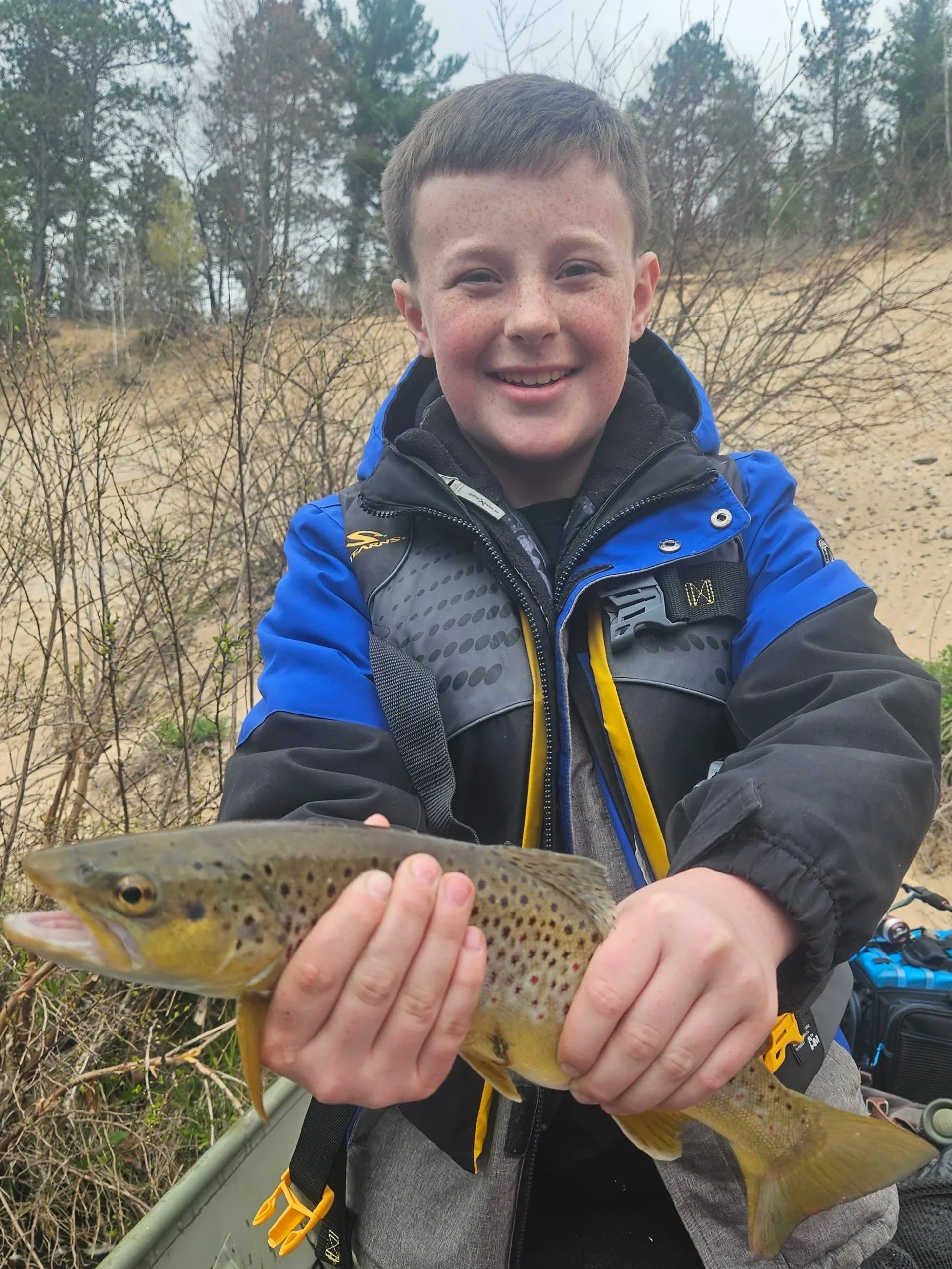 A smiling boy wearing a blue jacket holding a caught fish outdoors with trees in the background.