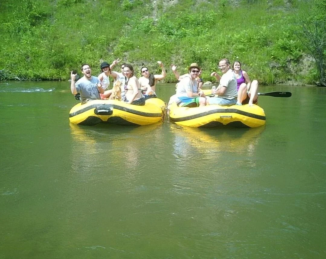 Group of people in two yellow inflatable boats on a river, smiling and waving at the camera, surrounded by green trees and foliage in the background.