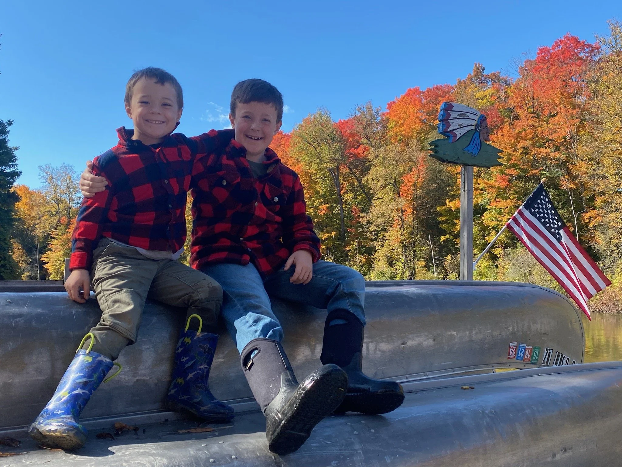 Two young boys in red and black plaid shirts sitting on the bow of a metal boat with an American flag and a decorative eagle figure, surrounded by trees with colorful autumn foliage, under a clear blue sky.