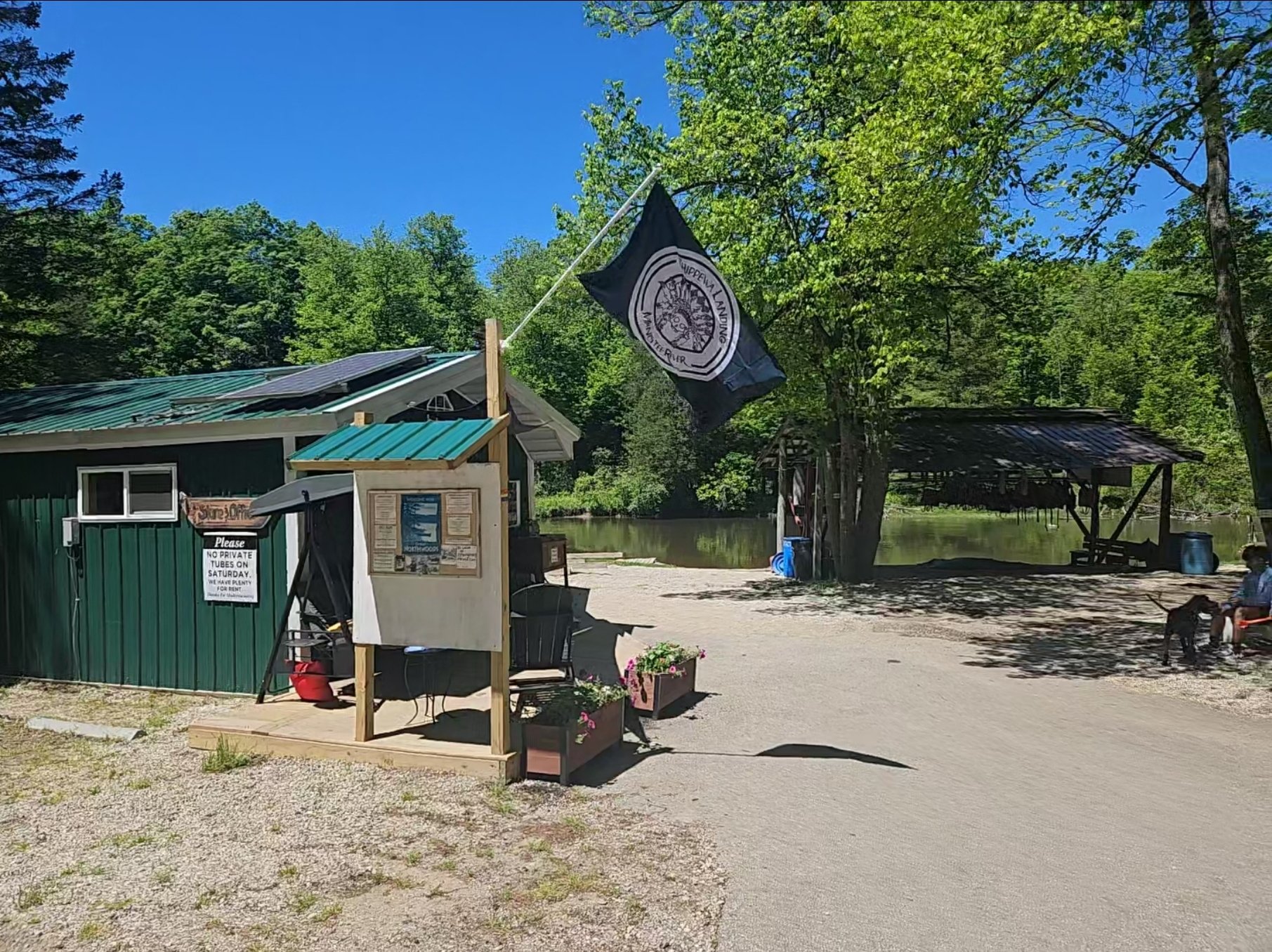 A lakeside outdoor area with a small green building, a flag with a logo and text saying "Upper Man Landings", a wooden bulletin board, potted flowers, and a shaded dock over the water surrounded by green trees and a clear blue sky.