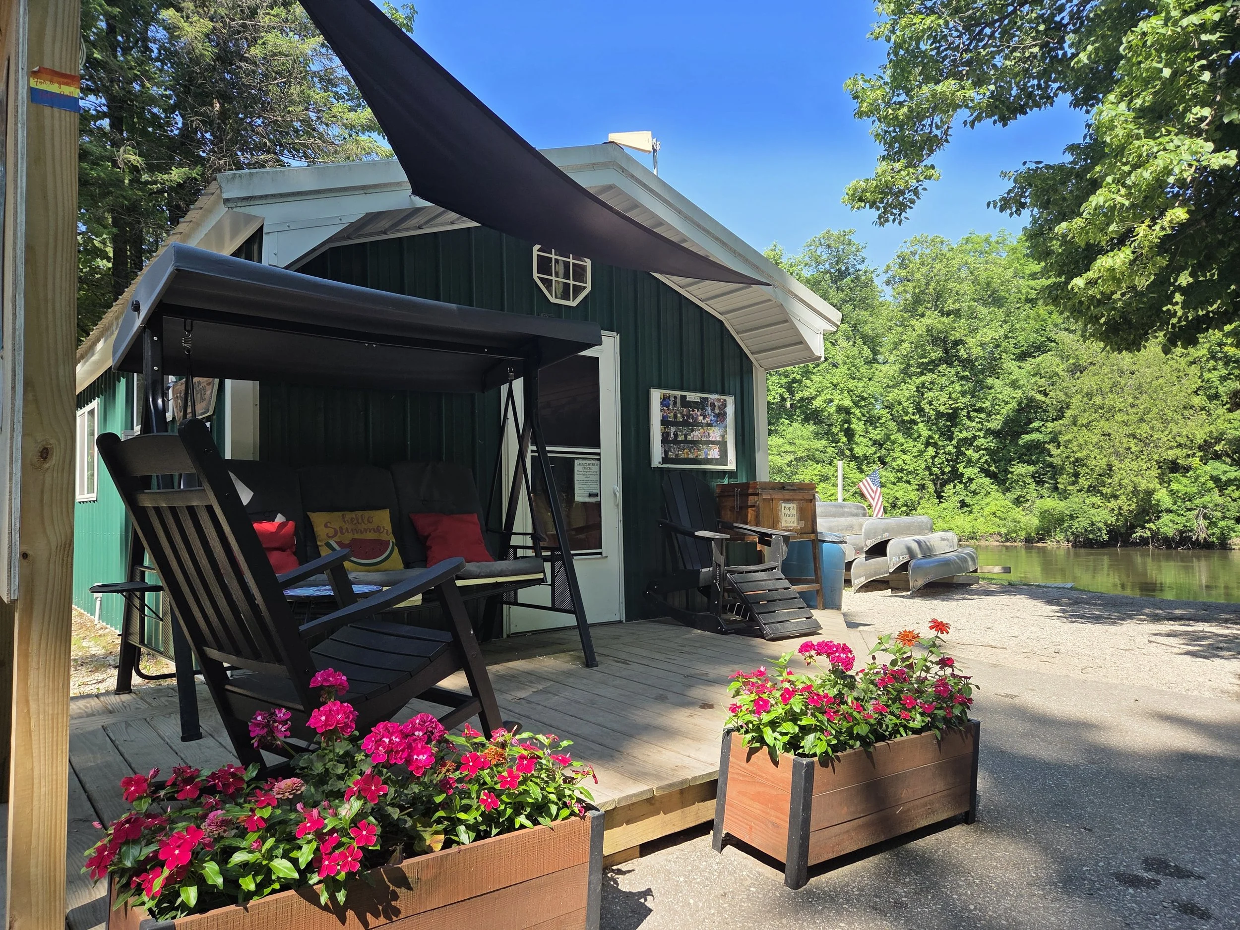 Outdoor scene featuring a deck with pink flowers in flower beds, a wooden swing with red and yellow pillows, and a green building with nautical flags, surrounded by lush green trees under a clear blue sky.