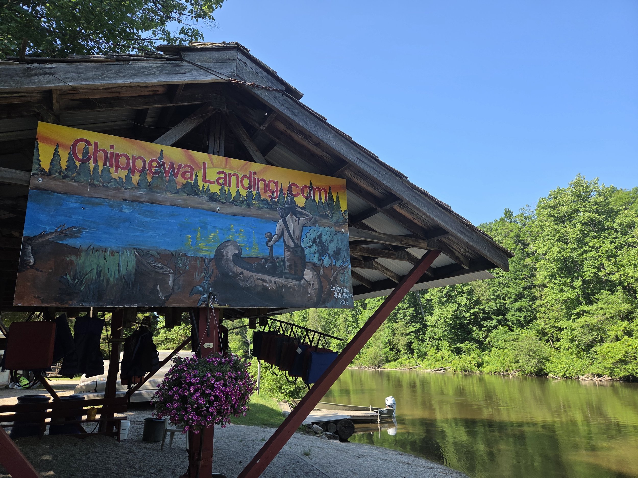 A colorful sign with the text 'ChippewaLanding.com' hanging under a rustic wooden roof near a river, with green trees, flowers, and a boat in the water.