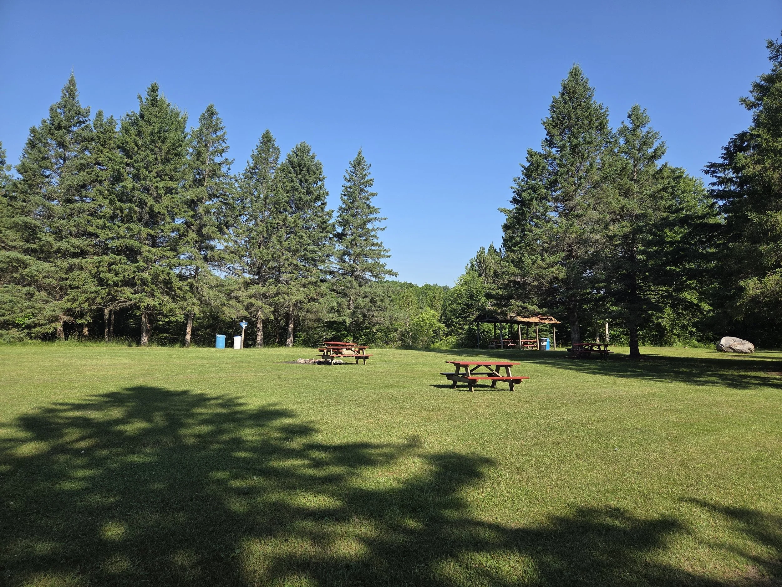 A grassy park area with picnic tables, a pavilion, and trees under a clear blue sky.