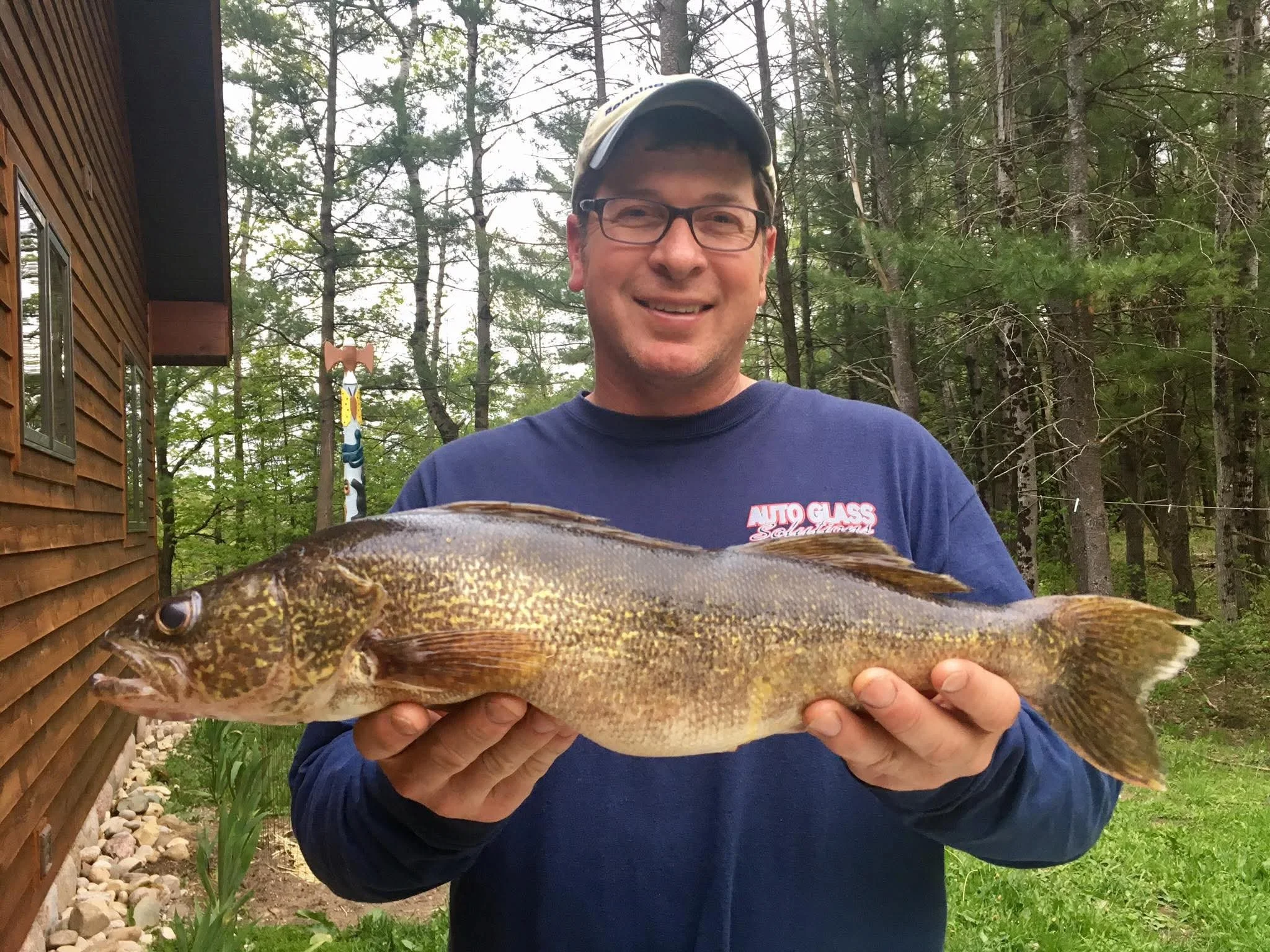 A man wearing glasses and a baseball cap is smiling while holding a large fish in front of him. The background shows trees and part of a building with wooden siding.