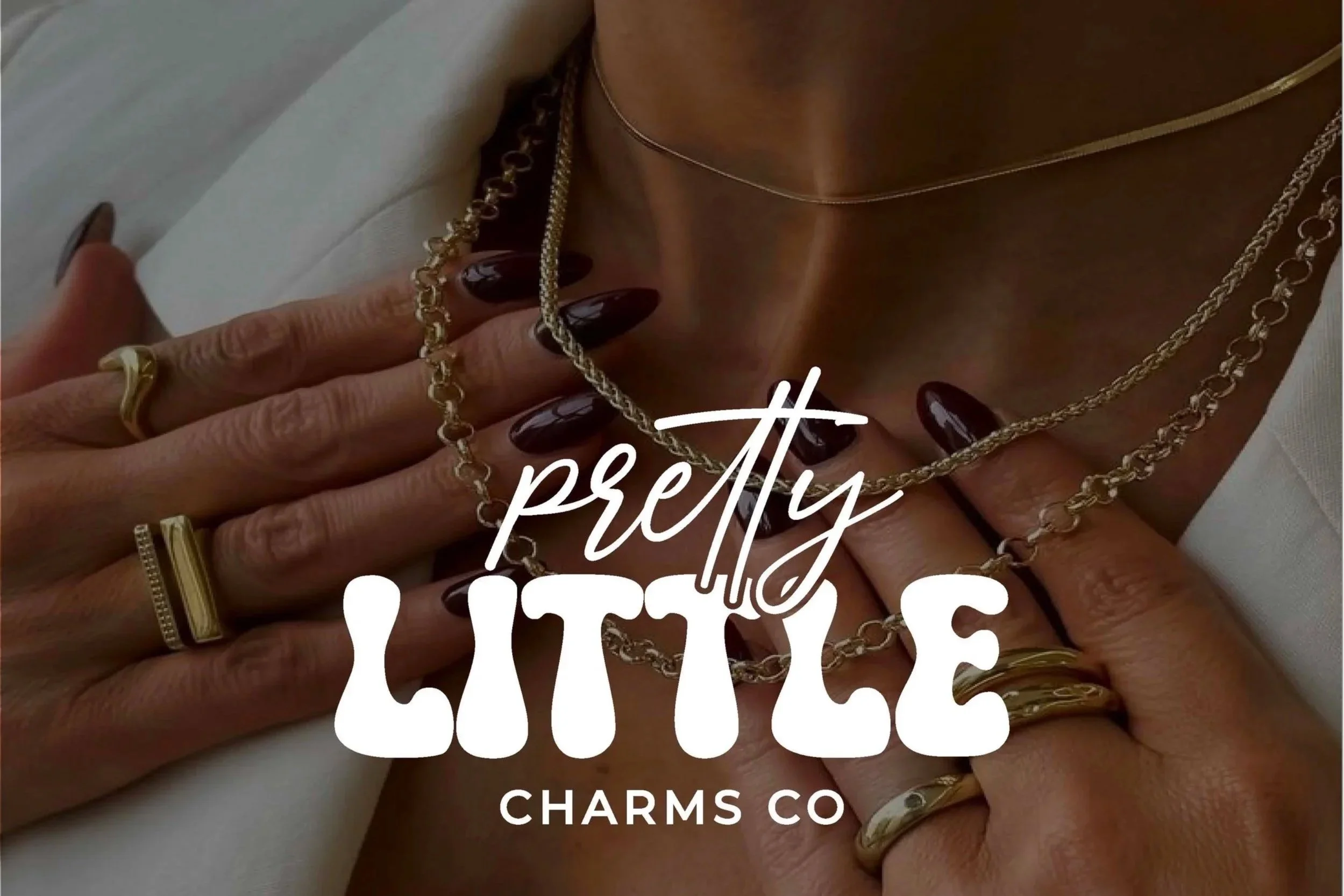 Close-up of a person's hand wearing gold rings and layered necklaces, with dark polished nails, and text overlay reading 'pretty' and 'Charms Co'.