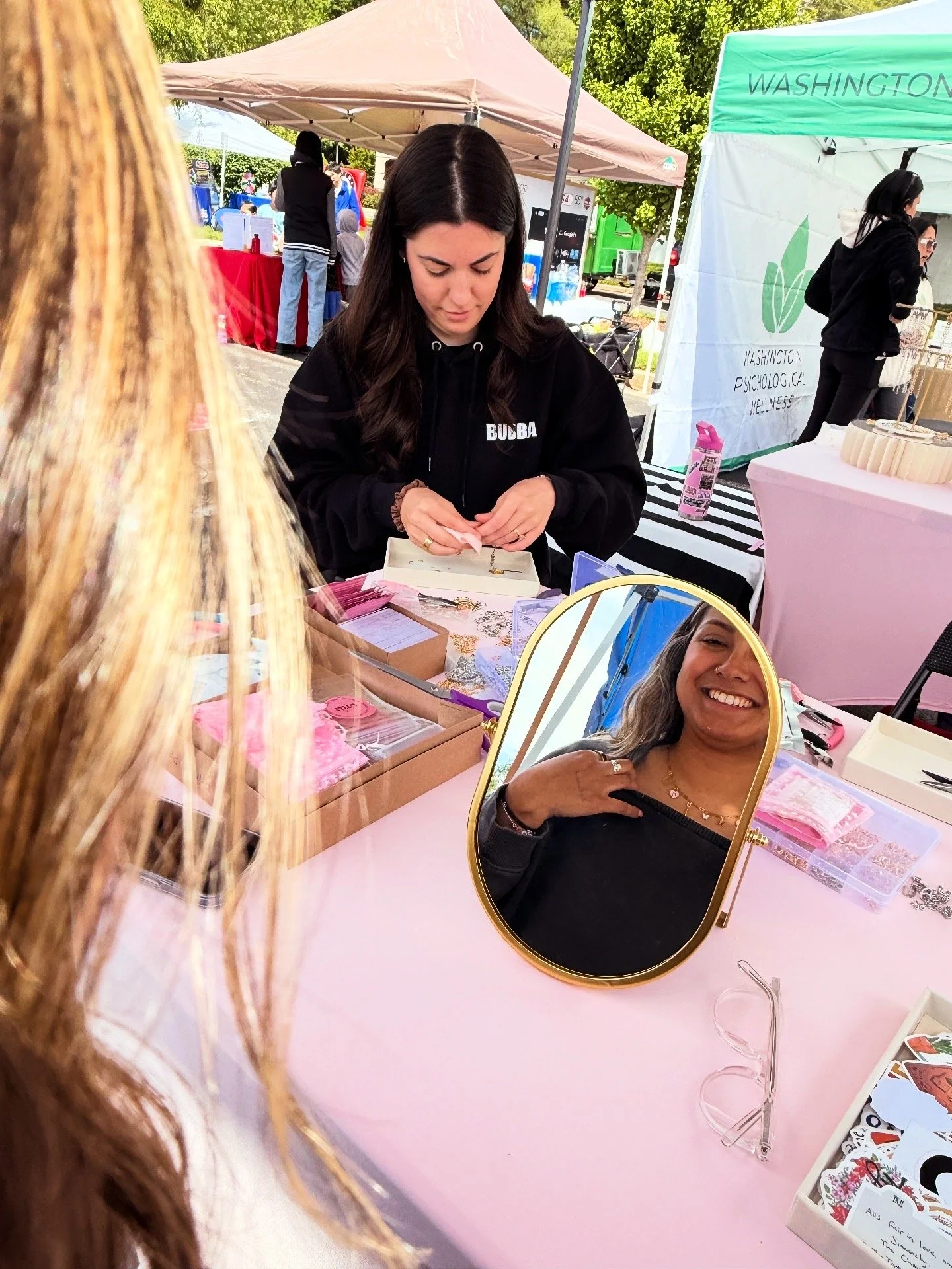 At a farmers market stall, a woman with long dark hair is sitting behind a table, jewelry and craft items, with a mirror showing her smiling face. Tents and people are visible in the background.