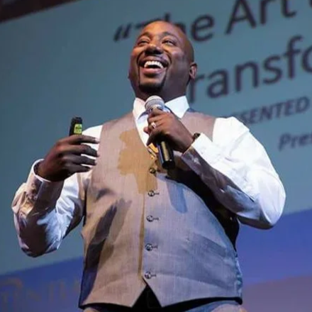A man in a vest holding a microphone and smiling, speaking at an event or presentation.