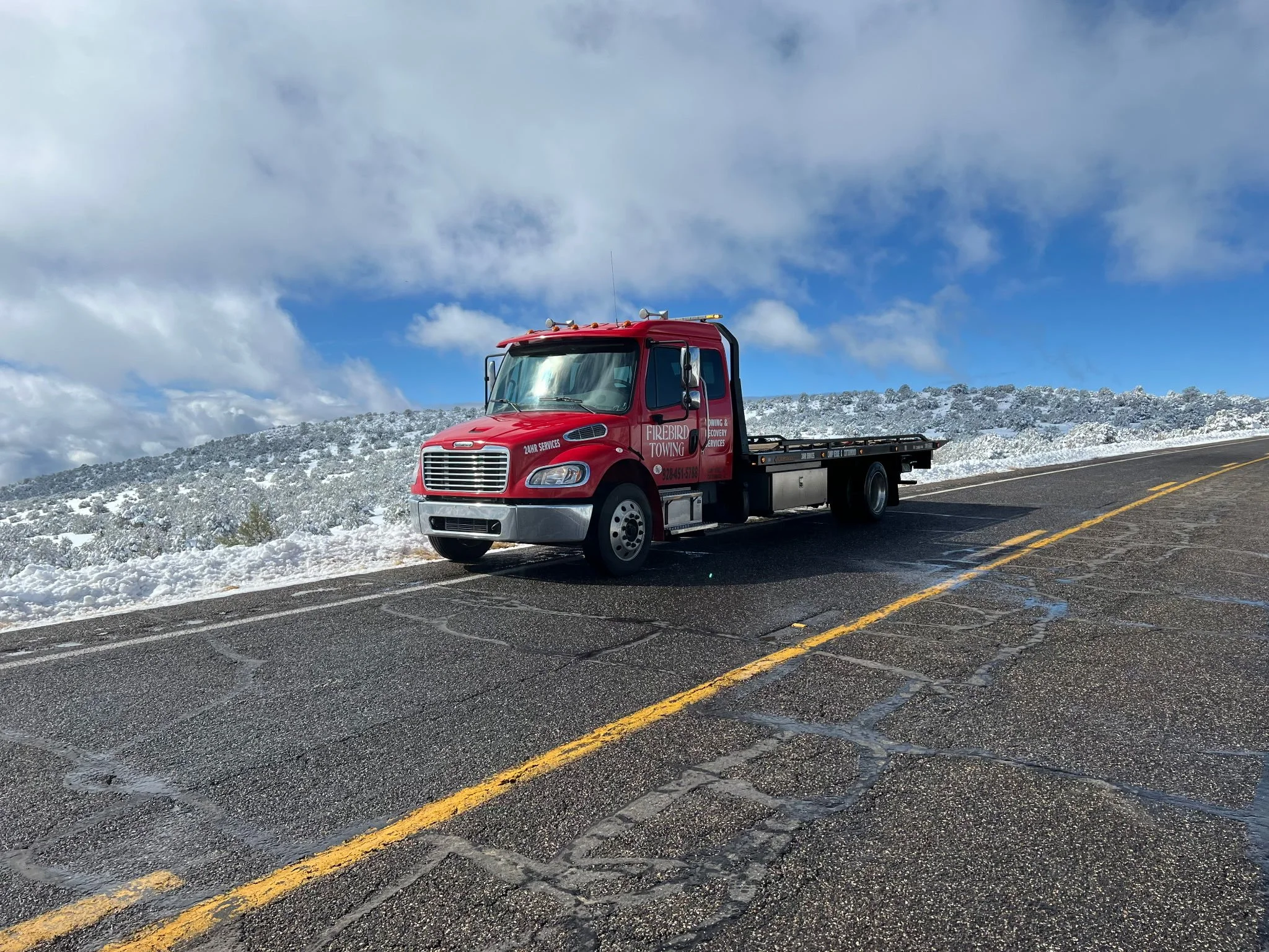 A red tow truck parked on a mountain road with snow-covered ground and a bright blue sky with clouds.