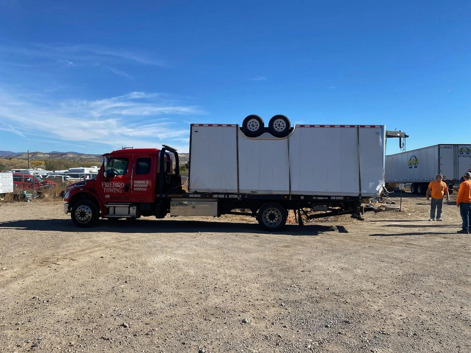 A red tow truck with a white cargo trailer attached, parked on a dirt lot. Two men in orange shirts are talking nearby, and other trucks are visible in the background under a clear blue sky.