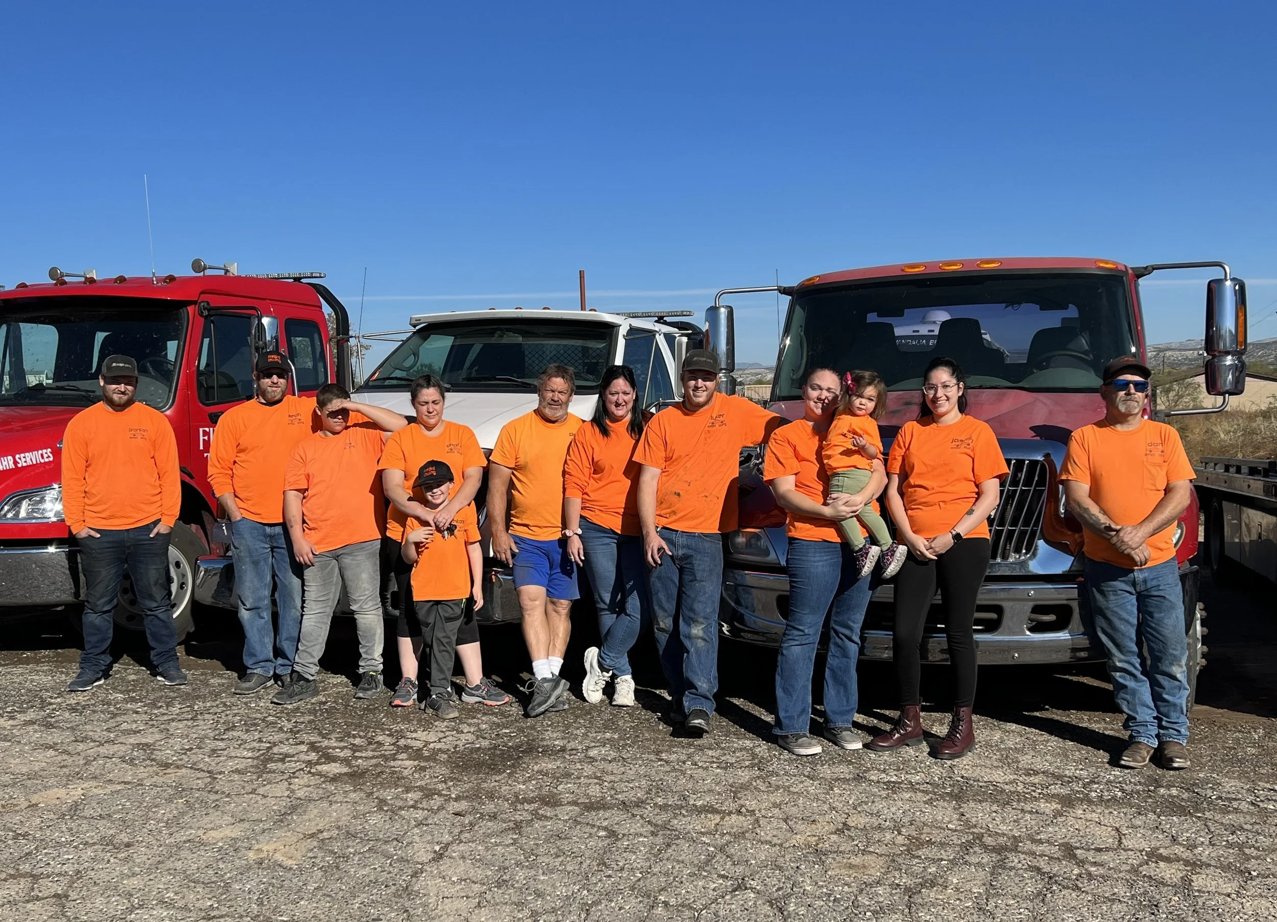 A group of people wearing matching orange T-shirts standing in front of three fire trucks, outdoors on a sunny day with a clear blue sky.