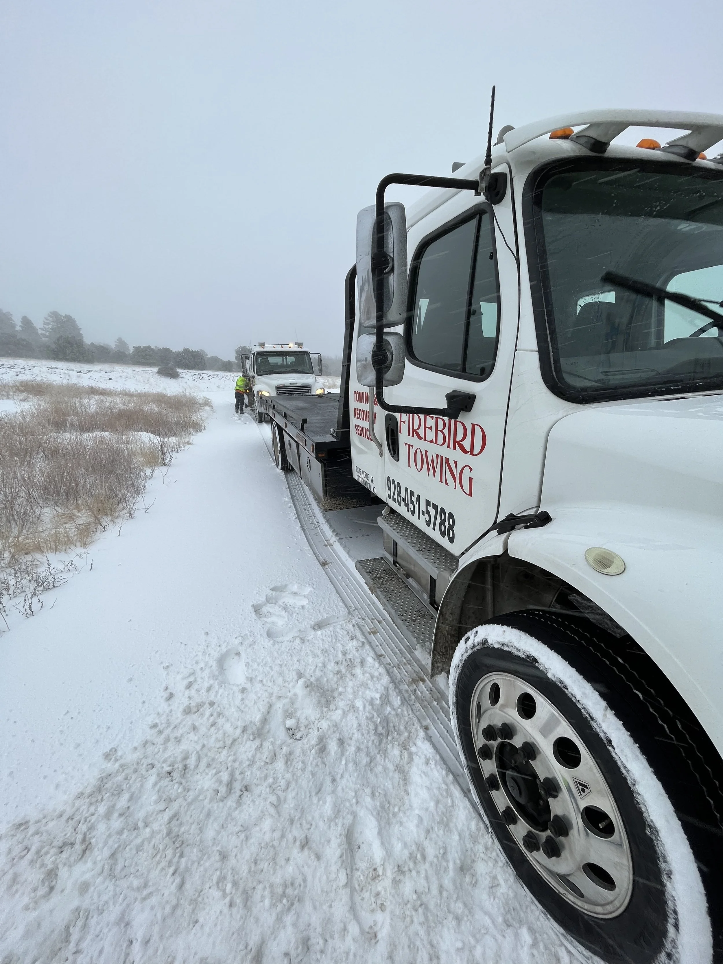 Two tow trucks on a snowy road, one in the foreground with a side mirror, and another in the background with a person in a yellow jacket standing nearby, with snow-covered grass and trees in the distance.