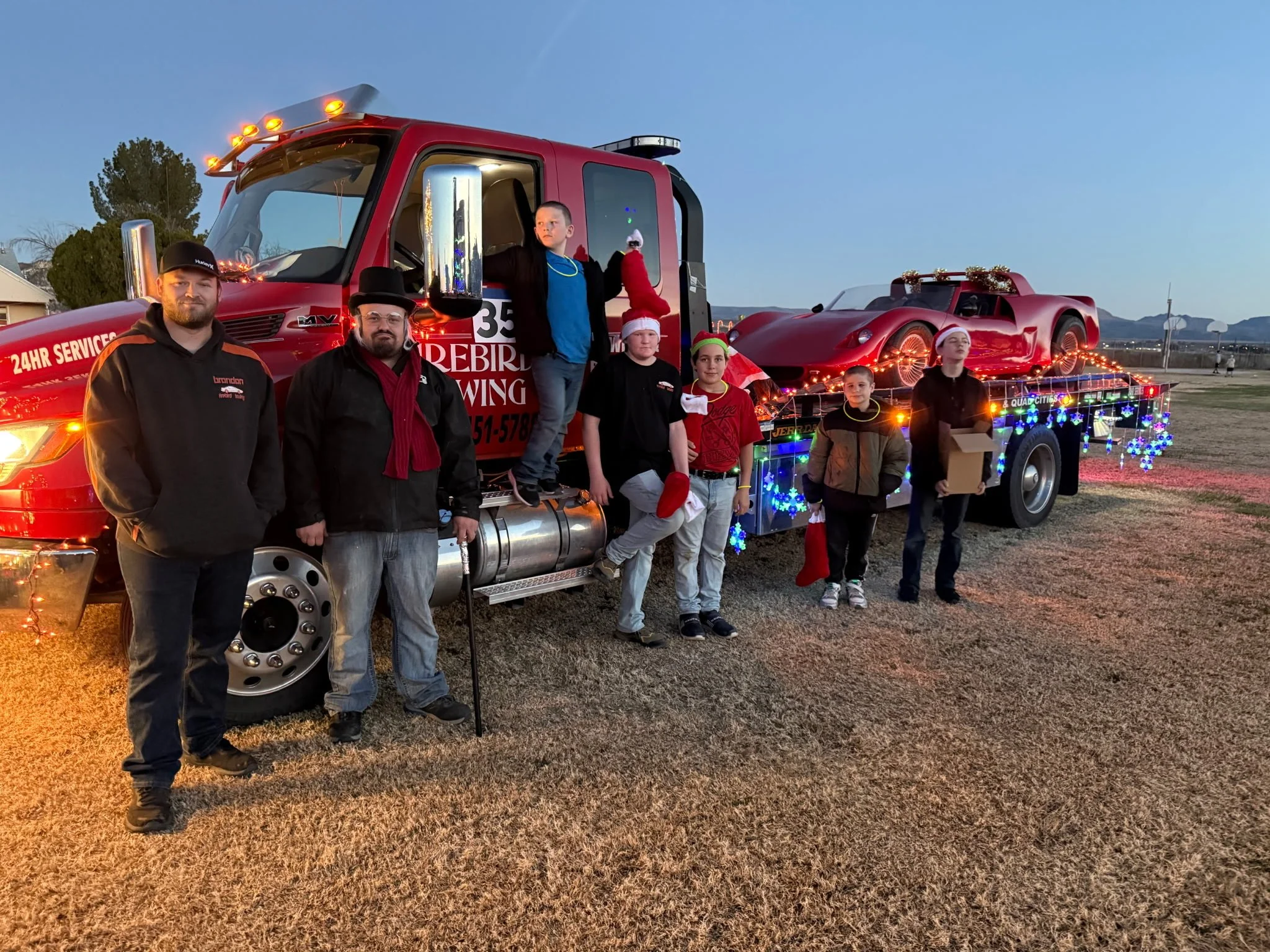 A group of people, including children and adults, stands in front of a decorated red fire truck with a sports car on a trailer, during a holiday event at dusk, with a clear sky and mountains in the background.