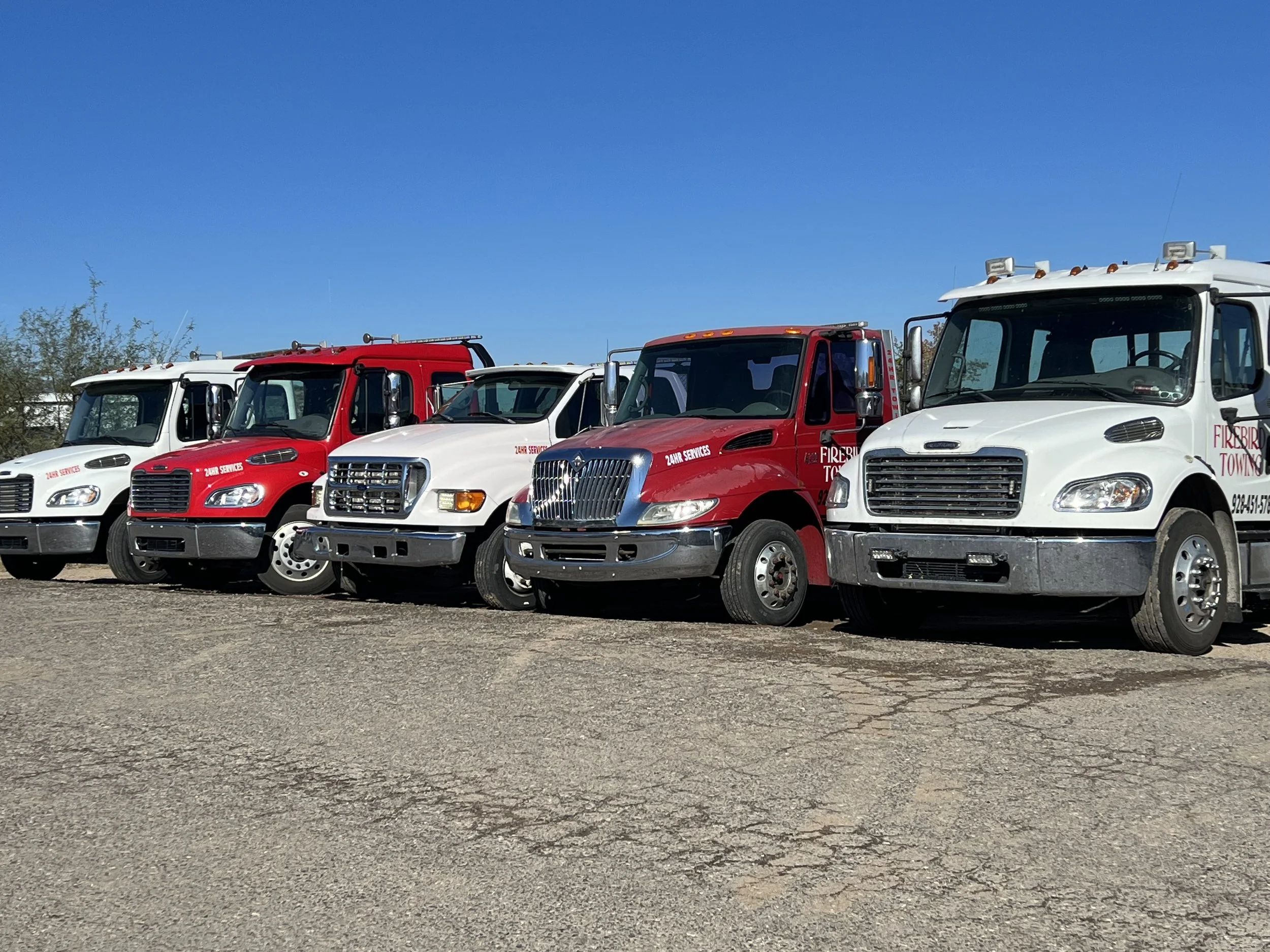 A lineup of fire department vehicles, including white and red trucks, parked on a gravel lot under a clear blue sky.