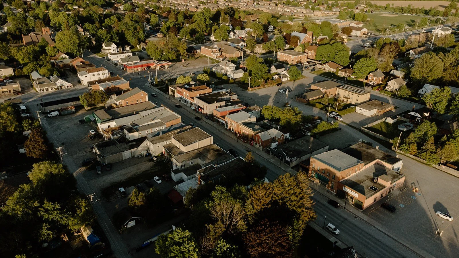 Aerial view of a small town with a main street lined with shops and businesses, residential houses surrounded by green trees, and parking lots, during daytime.