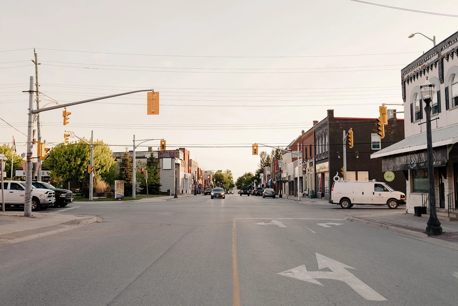 A quiet city street with parked cars, storefronts on both sides, traffic lights, and overhead power lines under a clear sky.