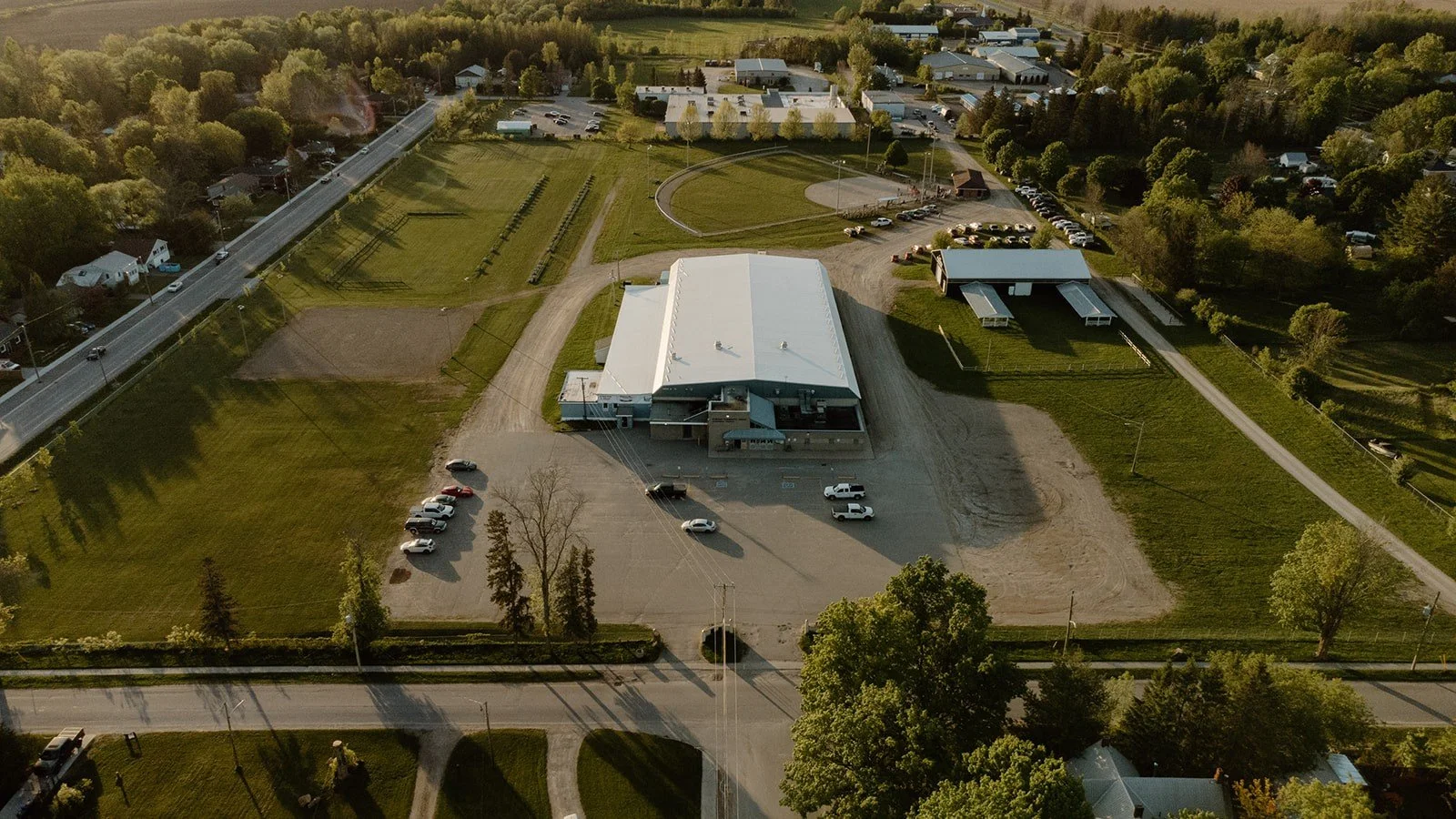 An aerial view of a large building with a white roof, surrounded by a parking lot with several cars, grass, and trees. There are additional structures, roads, and residential houses around this area.
